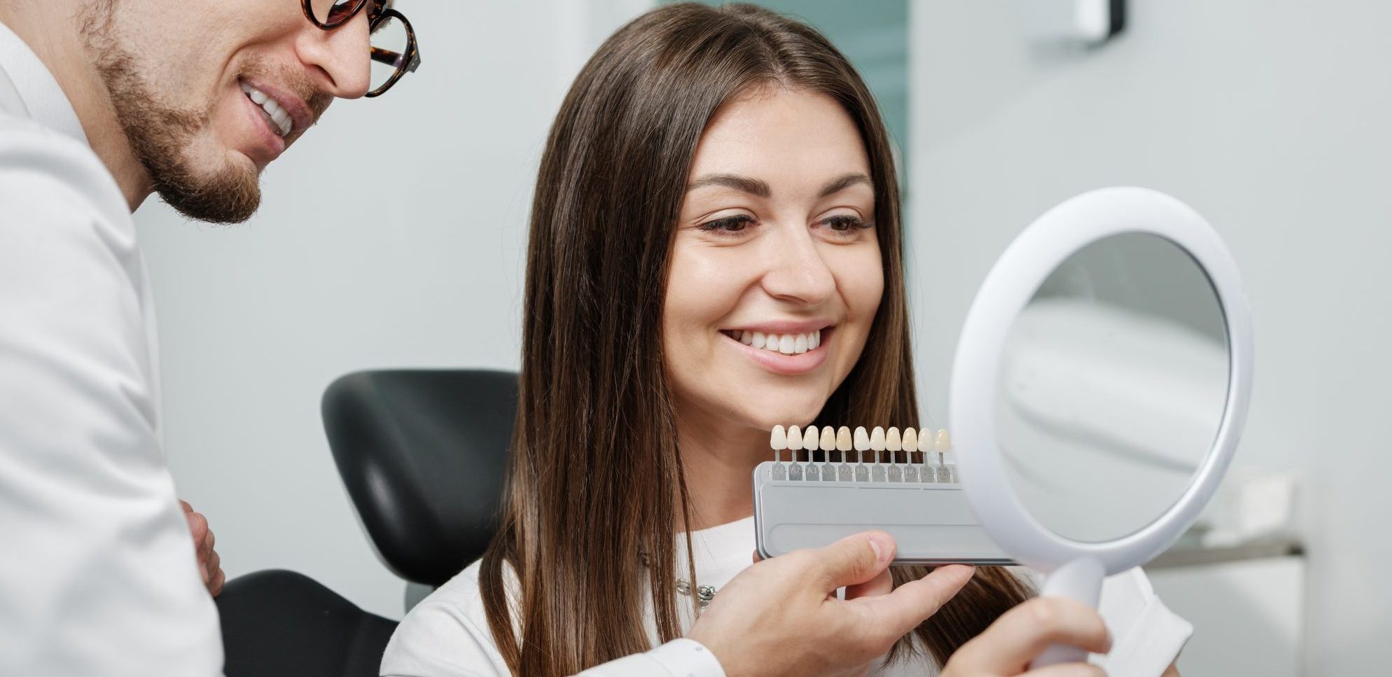 Young male dentist in white coat show veneers enamel color palette for beautiful patient woman sitting in a dental unit. Healthcare oral aesthetic treatment.