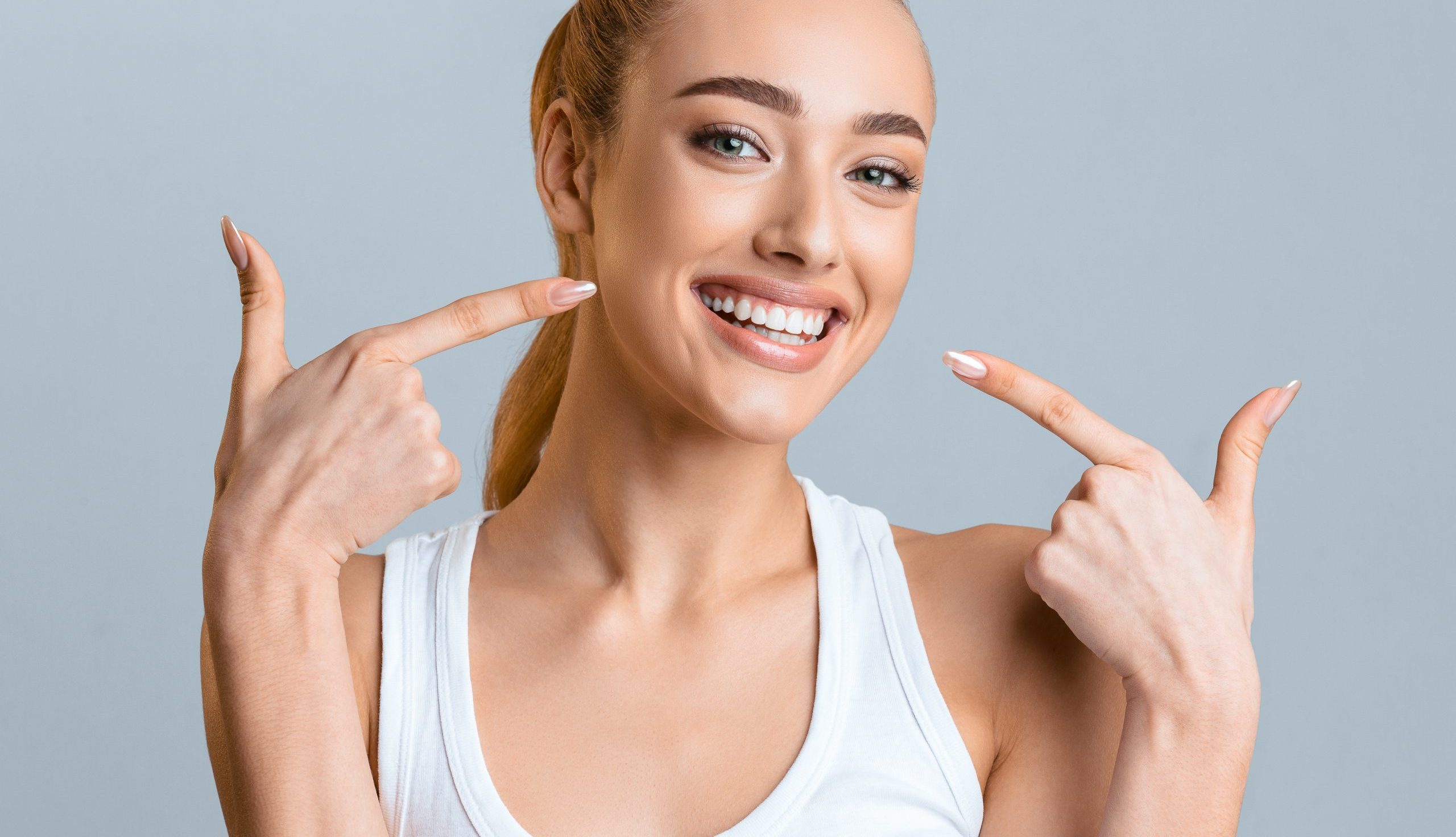 Orthodontic concept. Happy girl showing her beaming white teeth with two forefingers, grey background