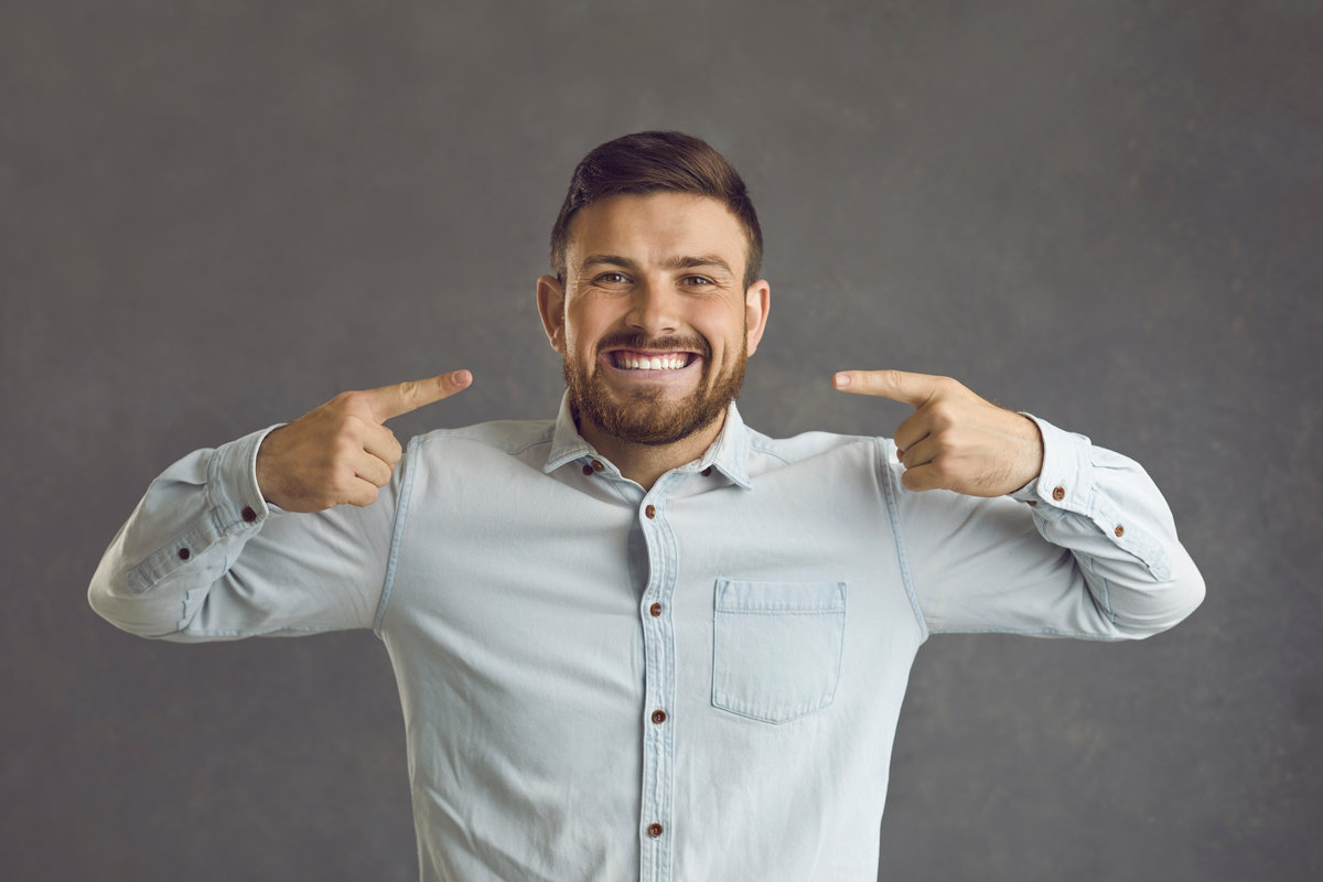 Portrait of an excited smiling client after visiting his dentist. Happy young man standing on a grey color studio background and pointing at his bright broad smile with perfect even white teeth