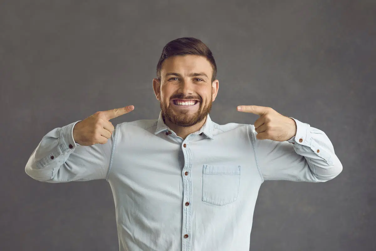 Portrait of an excited smiling client after visiting his dentist. Happy young man standing on a grey color studio background and pointing at his bright broad smile with perfect even white teeth