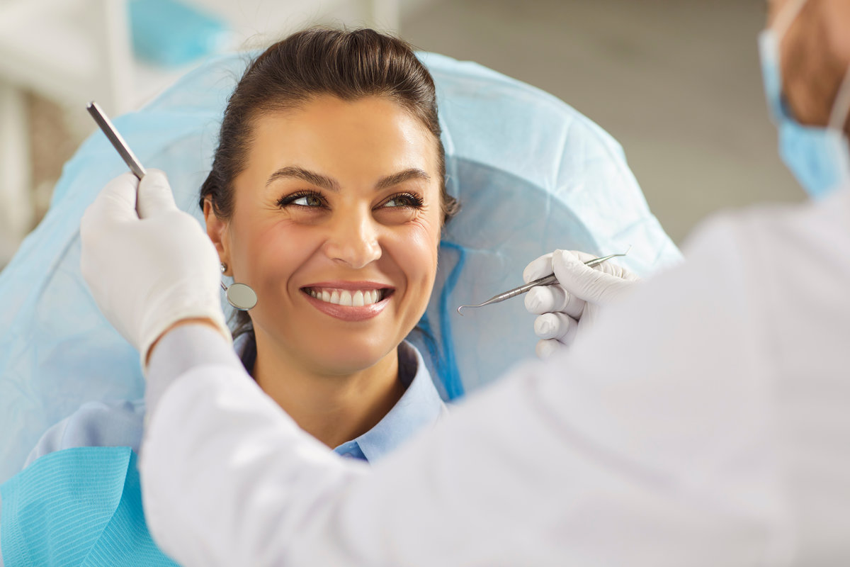 Happy woman at dental clinic receiving checkup with mirror and scaler, top view of dentists hands holding tools to check teeth. Female patient lying on chair, smiling, looking at doctor with trust