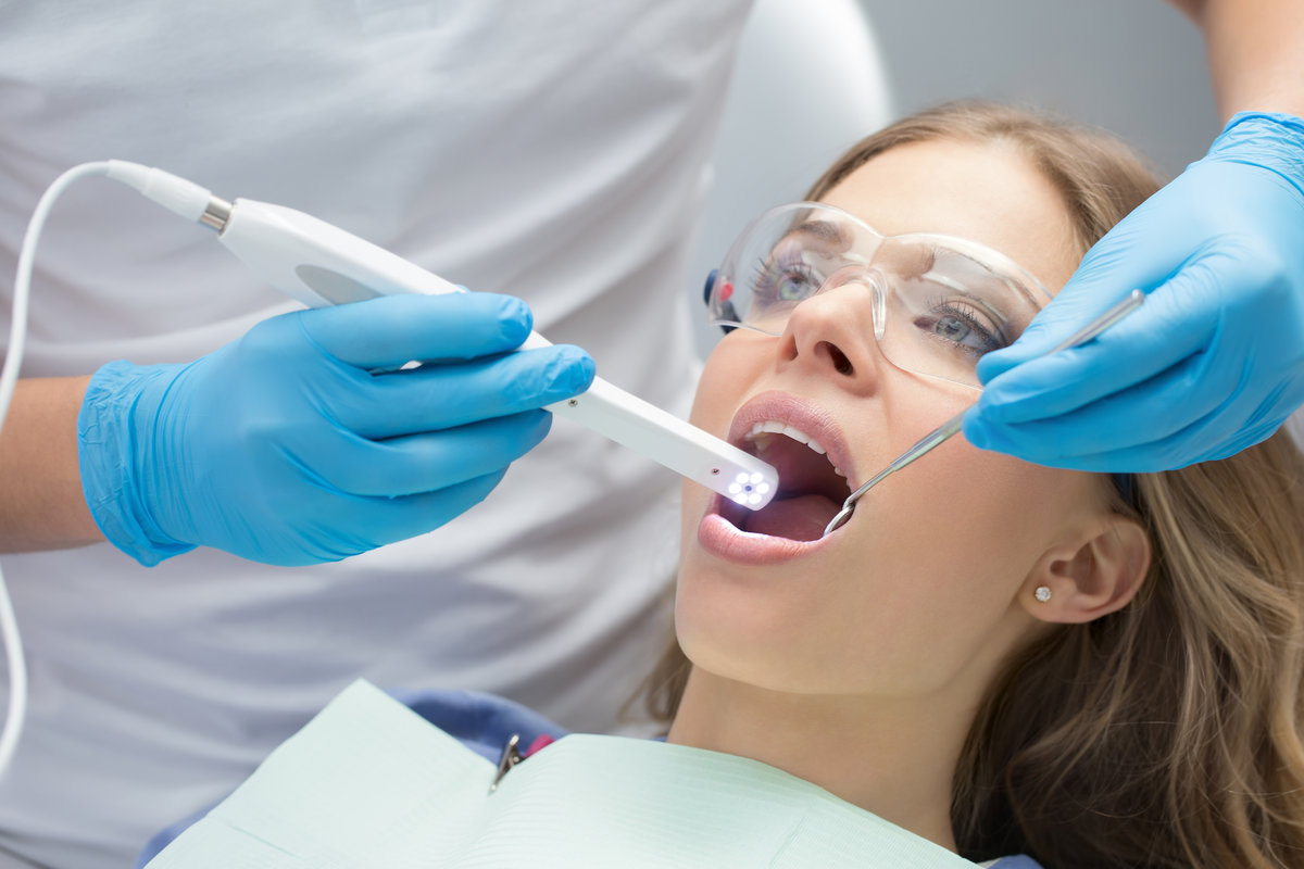 Lovely girl with opened mouth in patient bib and protective eyewear. Next to her there is a dentist in a white uniform with blue latex gloves. He diagnoses her teeth with a LED intraoral camera and a stem mouth mirror. Horizontal.