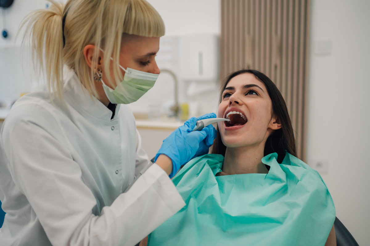 Female dentist wearing mask and gloves examining patient's teeth with dental intraoral camera in dental clinic, providing professional oral healthcare