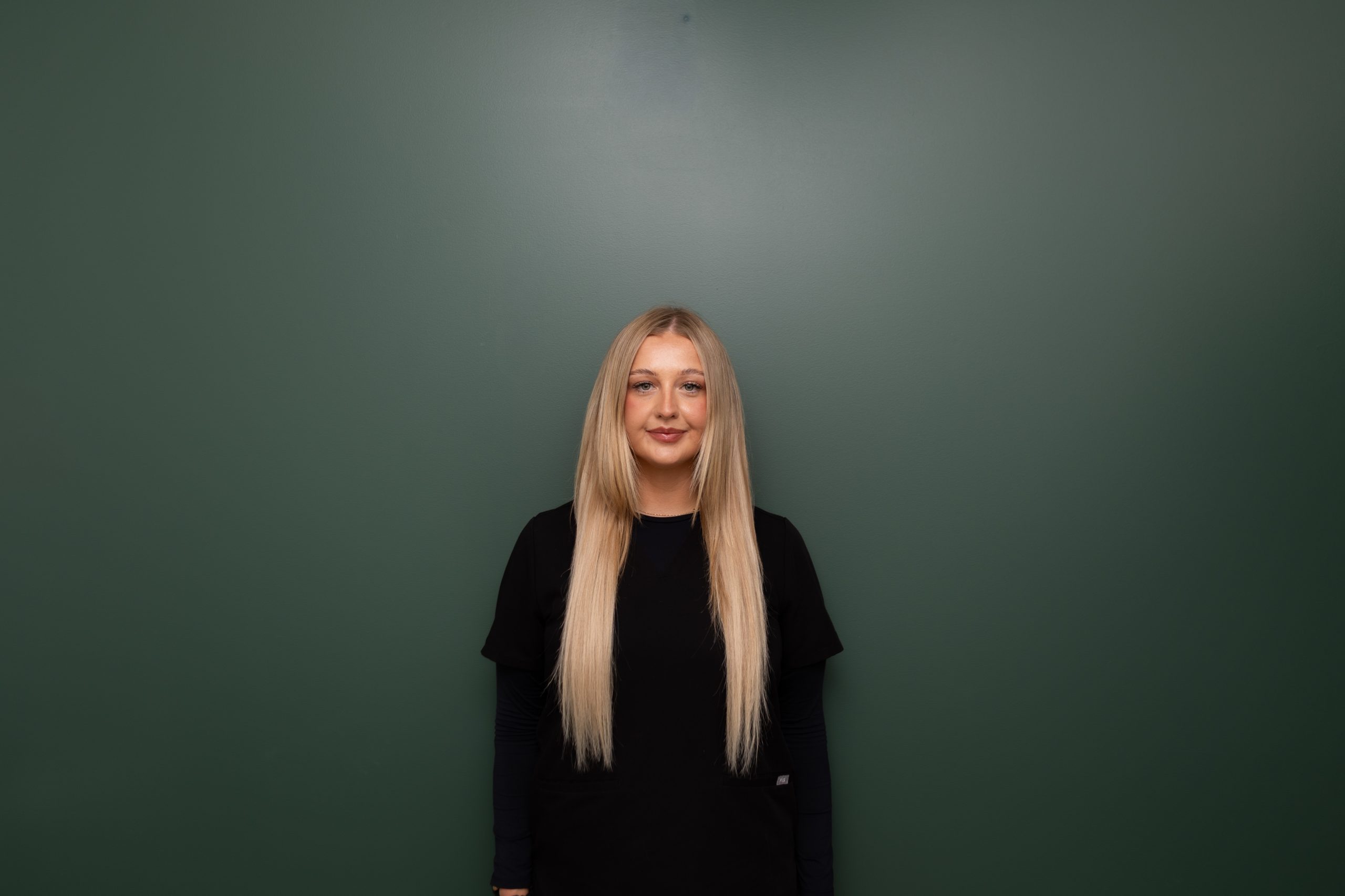 Woman with long blonde hair in black shirt smiling at camera against green background