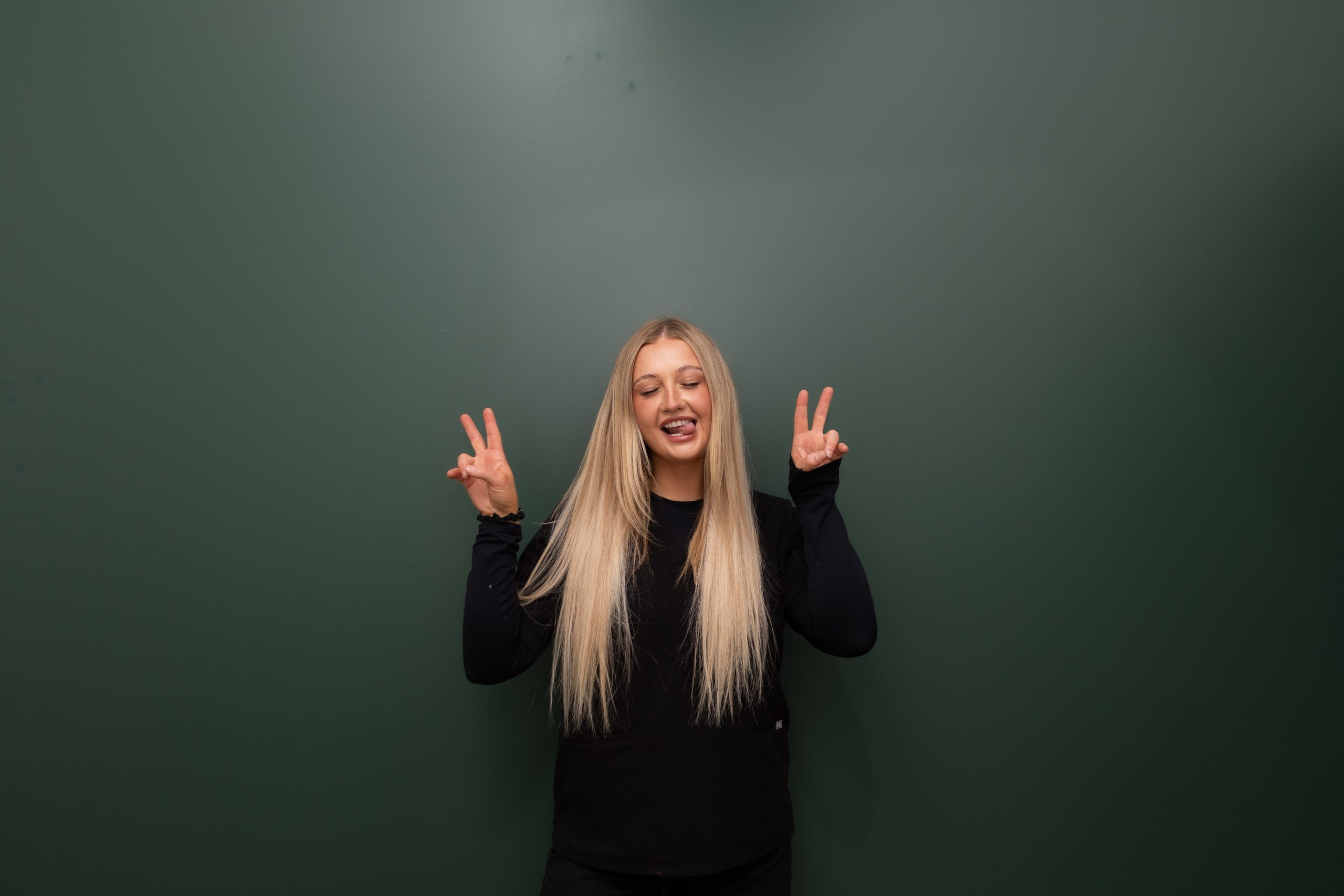 Woman with long blonde hair making peace signs against green background, eyes closed