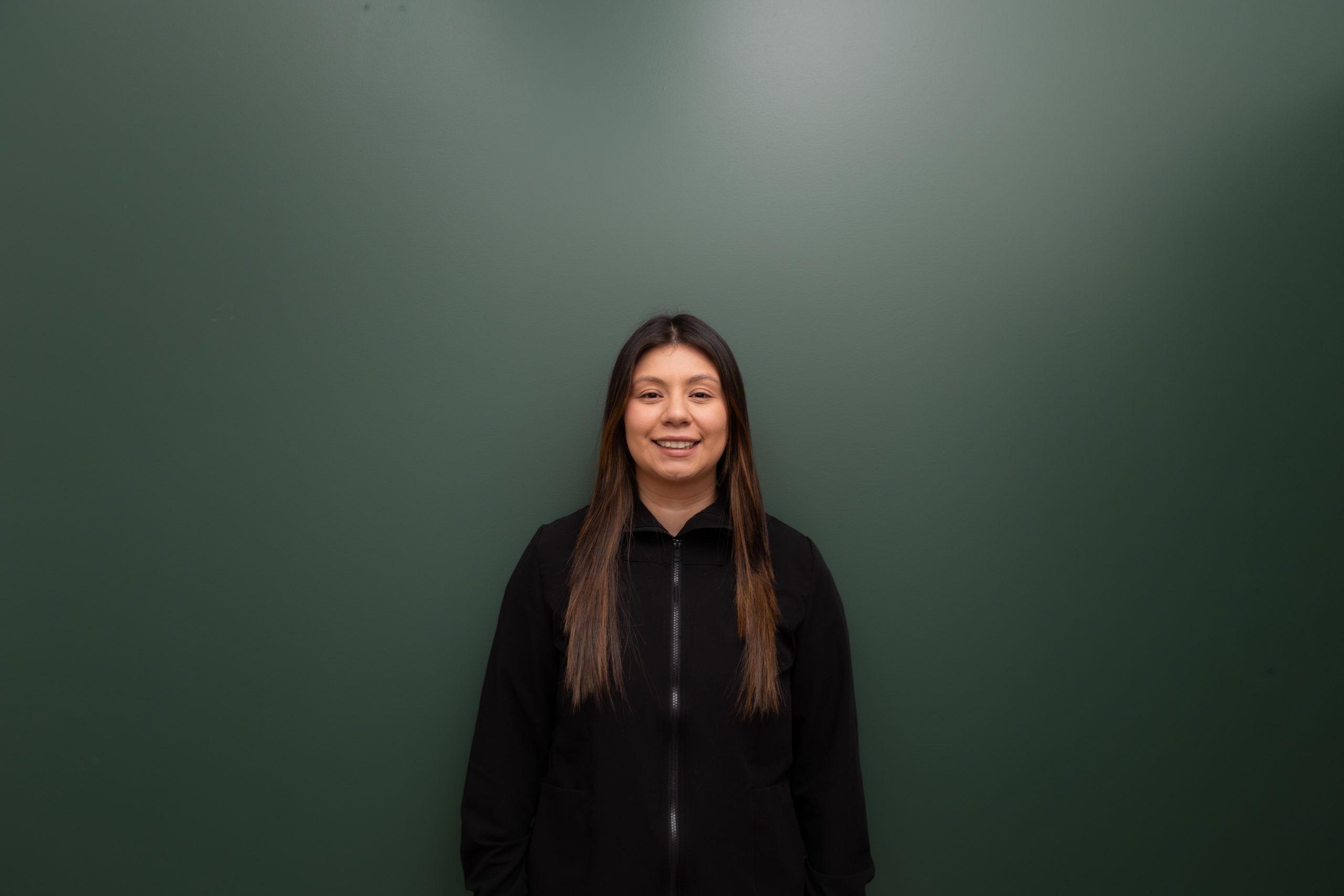 Woman with long dark hair in black jacket smiling at camera against green background
