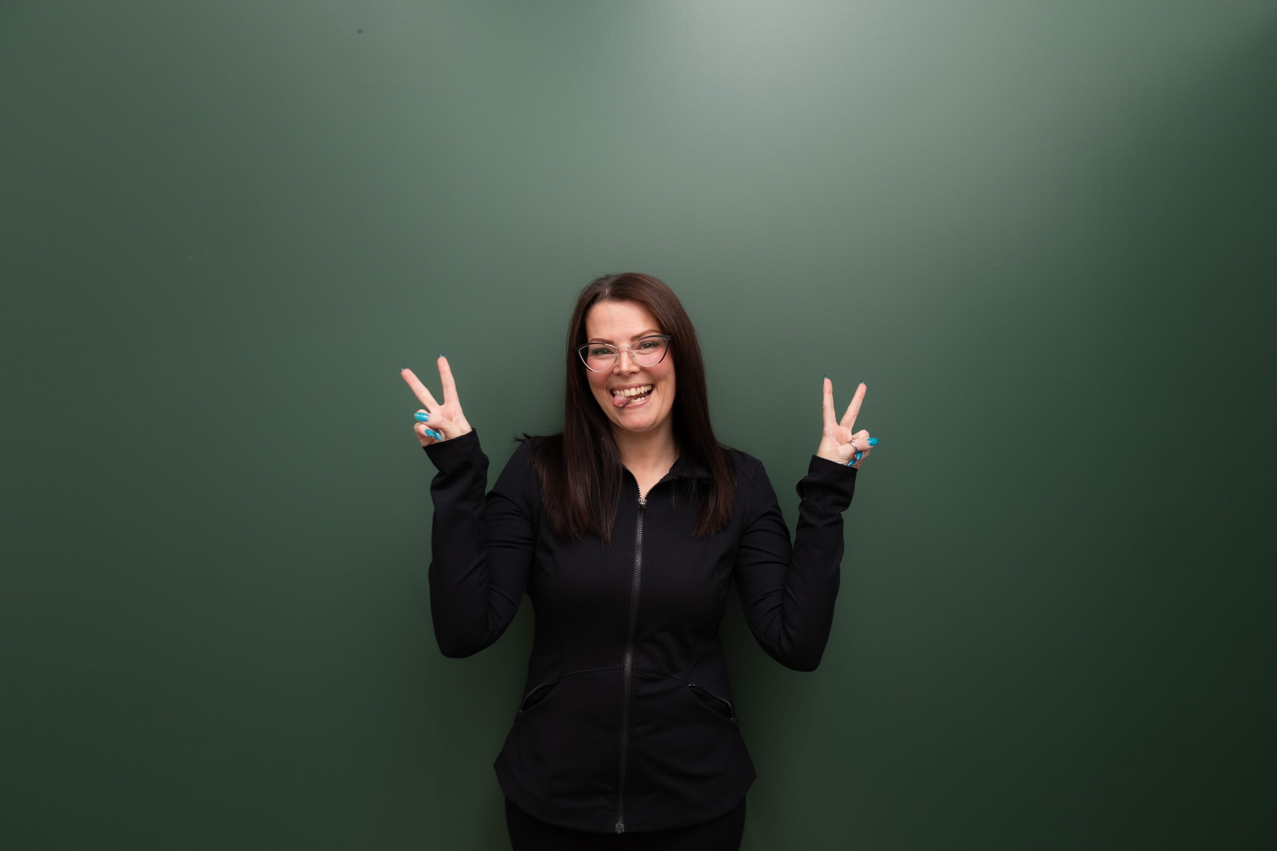 Woman in black jacket making peace signs against green background, smiling at camera