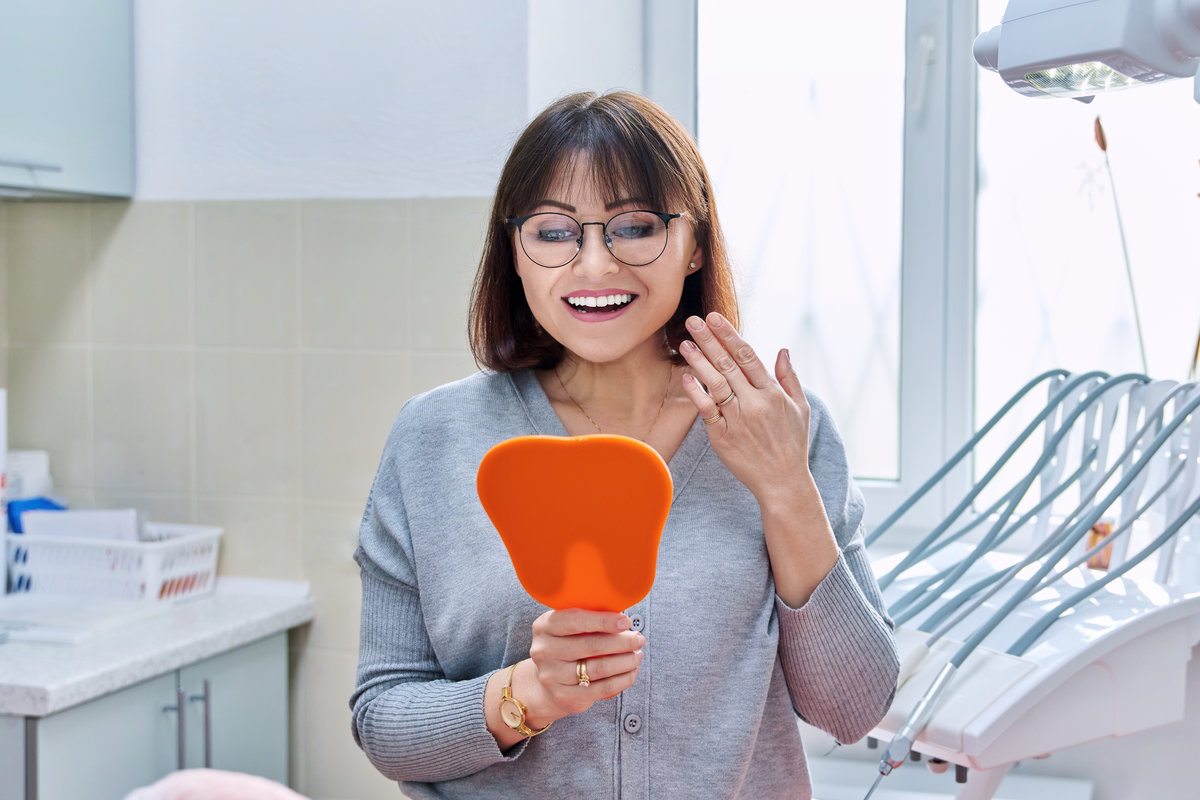 Happy middle aged woman dentist patient with a mirror in her hands looking at her teeth. Treatment, whitening, implants, medicine, health care and beauty smile concept