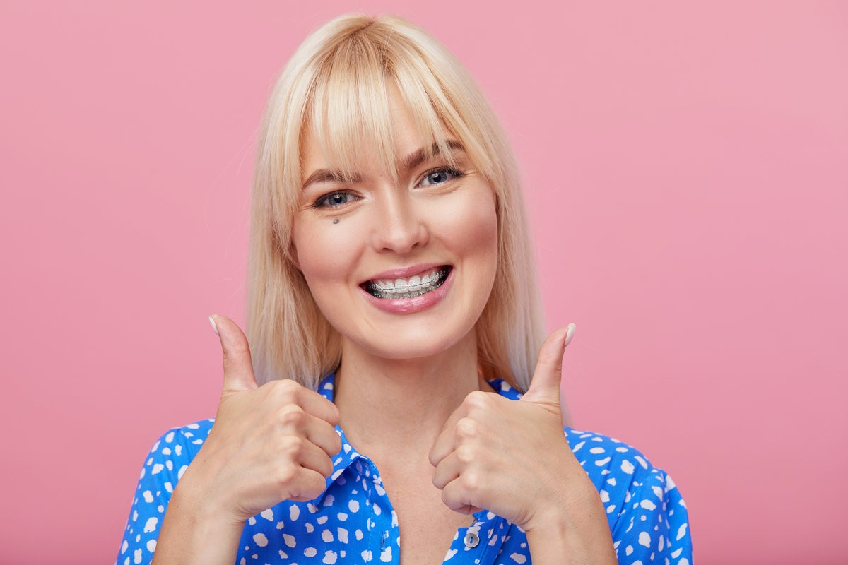 Cheerful happy young woman with blonde hair gesturing thumb up while pointing finger at braces on her teeth isolated over pink background.